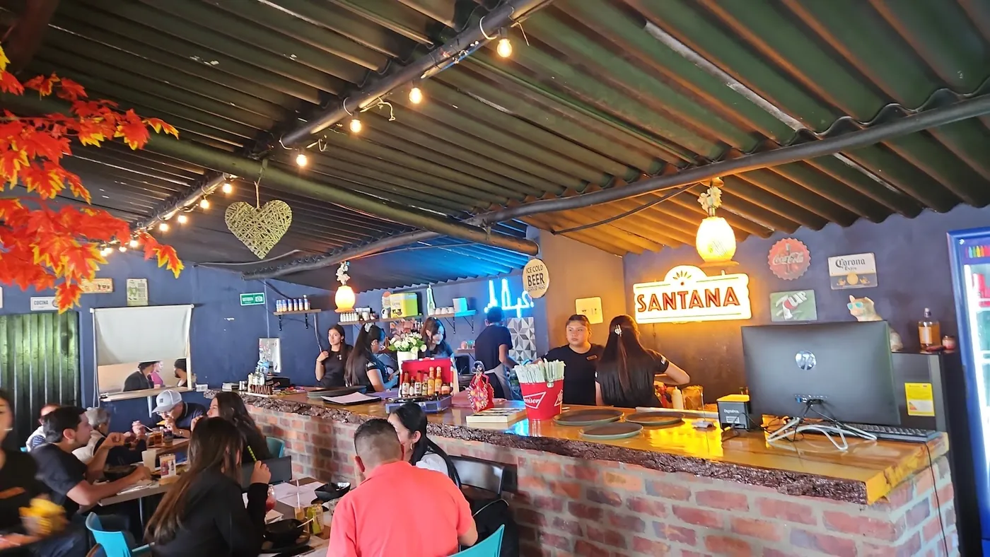 Terraza de Santana en Guasca: techo de teja, luces colgantes, sillas turquesa y el letrero neón de la casa